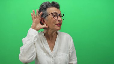 Gözlüklü, gri saçlı yaşlı bir kadın canlı yeşil bir arka plana karşı el pençe divan dinlerken beyaz bir gömlek giyiyor ve içten bir şekilde gülümsüyor..