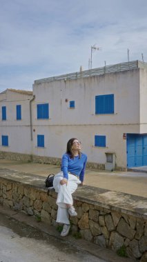 Geleneksel Akdeniz evinin önündeki taş duvarda oturan ve Mallorca 'da kıyı manzarasının keyfini çıkaran mavi panjurlu bir kadın.