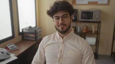 Ofis odasında gülümseyen sakallı genç adam, rahat ve kendinden emin bir havayı iletiyor, tipik ofis elemanları ile çevrili, raflar ve mikrodalga fırın gibi..