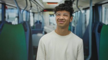 Gözlüklü ve beyaz kazaklı bir adam otobüs koridorunda yeşil koltuklu ve kabzalı gülümsüyor; iyimserlik.
