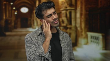 Gri ceketli genç adam süslü kilise binasının içinde yanak tutuyor; diş ağrısı rahatsızlığı endişesi.