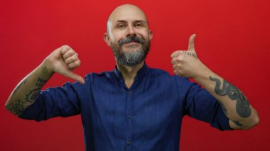 Sakallı kel bir adam, kırmızı arka planda onaylanmış ve onaylanmamış jestler yapıyor, mavi gömlek giyiyor, dövmeler yaptırıyor, yüz ifadelerinde karşıtlık tasvir ediyor..