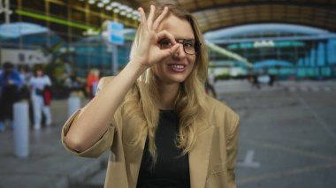 Genç sarışın kadın havalimanında parmaklarıyla hareket ediyor, canlı bir terminal ortamında gülümsüyor, gözlük ve bej ceket takıyor, seyahat ve macera hissini yakalıyor..