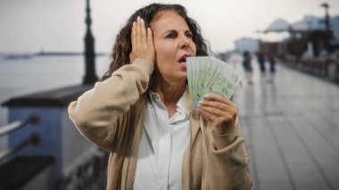 Singapur 'da, deniz kenarında orta yaşlı bir kadın, kalabalık bir liman bölgesinin yakınındaki gürültüden rahatsız olduğunu belirten bir ifadeyle, elinde Singapur doları tutuyordu..