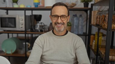 Kahve makinesi ve bulaşıklarla dolu raflarla çevrili bir ofiste gülümseyen, samimi ve profesyonel bir ortam sergileyen olgun İspanyol bir adam..