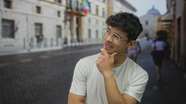 Молодой латиноамериканец в очках, рука на подбородке, думая на мощеной солнцем улице в историческом центре города; любопытство.