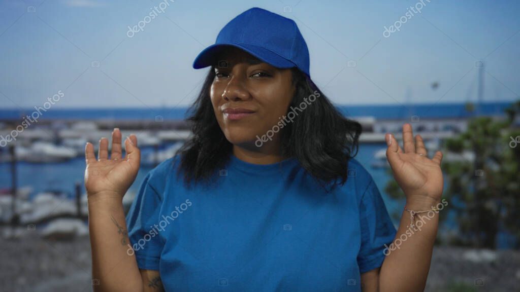 Blue cap and shirted courier woman raises both hands on a street outdoors beside harbor boats; frustration.