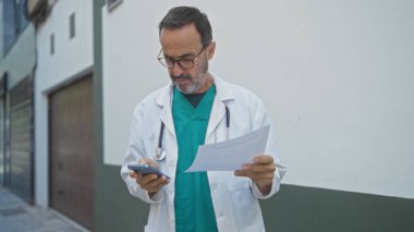 Tıbbi mantolu İspanyol bir adam, şehir sokaklarında doküman okurken ve telefonunu kontrol ederken, hastane ya da kliniğin yakınında profesyonel bir ortam izlenimi uyandırıyor..