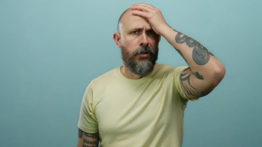Sakallı ve dövmeli bir adam mavi bir duvar arkasında kafası karışmış, soluk bir gömlek giyerken kafasına dokunuyor..
