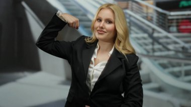 Alışveriş merkezindeki genç sarışın kadın iş kıyafetleriyle kol gücünü arttırıyor. Yürüyen merdivenlerle şehir merkezindeki bir alışveriş merkezine karşı güven ve başarı gösteriyor..