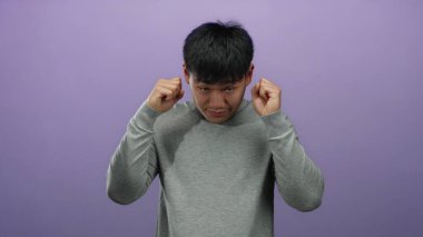 Gri kazaklı genç adam, yorgun ya da stresli görünen mor arka plana gözlerini sürterek, minimalist bir ortamda yorgunluk ya da endişeye işaret ediyor..