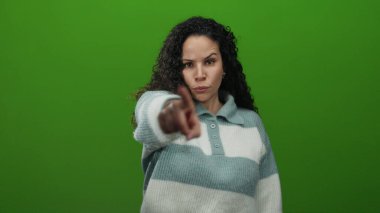 Yeşil arka plana karşı davetkar bir jest yapan, çizgili süveter giyen, iletişimi ifade eden, İspanyol kökenli, canlı bir ortamda izole edilmiş, genç görünümlü bir kadın..