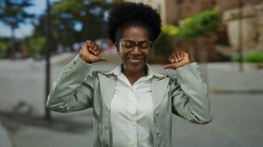 Gözlüklü bir kadın, yeşil bir caddede kendine güveni tam bir şekilde kendini gösteriyor. Açık renk bir ceket giyiyor ve hareketleriyle neşe saçıyor..