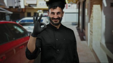 Sakallı İspanyol şef gülümsüyor ve açık havada güneşli bir sokakta siyah üniformalı jest yapıyor..