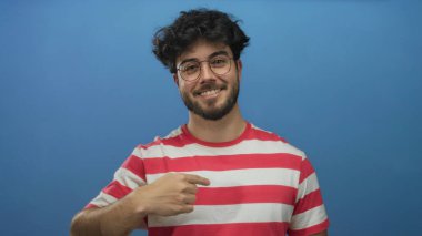 Çizgili gömlek giyen, sakallı ve gözlüklü genç İspanyol bir adam kendini işaret ediyor ve sağlam mavi bir arka plana karşı baş parmak işareti yapıyor..