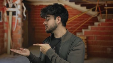 İnşaat alanının içinde bitmemiş tuğla duvarlar ve görünen kablolarla avucunun içini gösteren sakallı İspanyol bir adam..