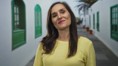 Sarı kazaklı, uzun saçlı bir kadın yeşil pencereli büyüleyici caddenin yanında kendine güvenle gülümsüyor, canlı, güneşli bir ortam yaratıyor..