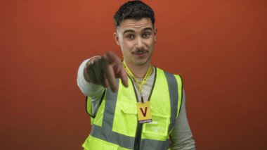 Yansıtıcı yelek giyen genç İspanyol adam izole edilmiş turuncu arka plana karşı gönüllü olarak işaret ediyor ve dost ifadesiyle alkışlıyor..