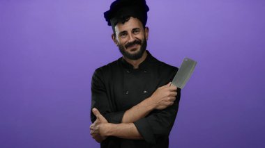Sakallı bir şef mor arka planda duruyor. Kollarını kavuşturmuş, kendine güveni ve profesyonelliği parlak siyah üniformasıyla yayıyor..
