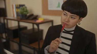 Çizgili kazak giyen ve ofis ortamında kalemle kafa yoran genç bir kadın. Kitaplar, bitkiler ve organizasyonel dekorlar içeriyor..