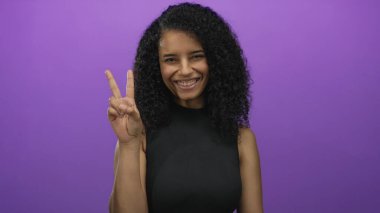 İspanyol kadın gülümsüyor ve mor arka plana karşı barış işareti koyuyor, canlı bir kişilik sergilerken pozitiflik ve kendine güven yayıyor..