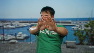 Üzerinde gönüllü yazan yeşil gömlekli genç adam açık mavi gökyüzünün altında bulanık teknelerle sahil limanında yüzünü kapatıyor.