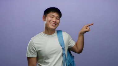 Mavi sırt çantalı beyaz gömlekli genç adam heyecanlı bir şekilde izole edilmiş mor arka planı işaret ediyor.