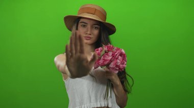 Hasır şapka giyen genç İspanyol kadın yeşil bir stüdyoda el hareketi çekerken elinde pembe bir buket tutuyor..