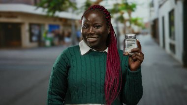 Afrika kökenli Amerikalı bir kadın, elinde bir kavanozla kalabalık bir caddede yeşil bir kazak giyiyor ve düşünceli bir ifade sergiliyor..