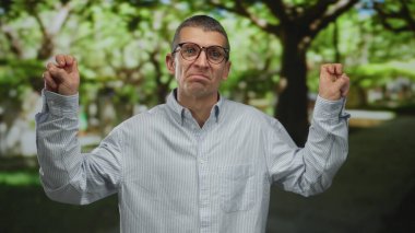 Parkta gözlüklü bir adam hava tırnak içinde iğneleyici sözler sarf ediyor ve açık havada yemyeşil bir ortamda günlük kıyafetler giyiyor..