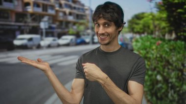 Bulanık trafik arkaplanı ve kaldırım çizgilerine karşı güneşli şehir caddesinde boş kaldırıma doğru avuç içi açık jestler yapan genç adam;.