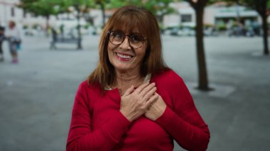 Yaşlı İspanyol kadın göğsünde gözlüklerle gülümsüyor ve şehir sokaklarında kırmızı süveter giyiyor. Ağaçlar bulanık ve arka planda insanlar var..