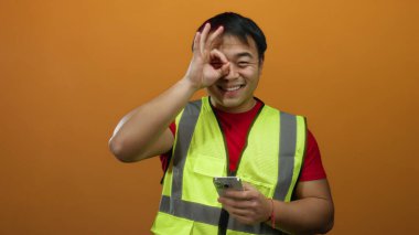 Adam gülümsüyor ve turuncu arka planda yüksek görünürlüklü yelek giyiyor.