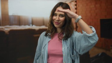 Bir inşaat alanında gülümseyen genç İspanyol kadın, pembe bir gömlek ve kot ceket giyiyor, arka planda tuğla duvarlarla kapalı bir yerde duruyor, sıcaklık ve pozitiflik taşıyor..