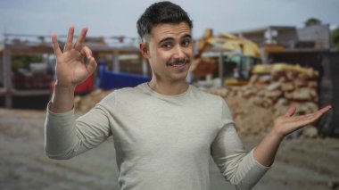 İnşaat alanındaki genç İspanyol adam elleriyle iyi bir jest yapıyor ve makinelerin arasında güvenle gülümsüyor..