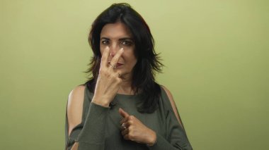 Orta yaşlı bir kadın stüdyoda kamerayı doğrudan zeytin yeşili duvara doğrultuyor..