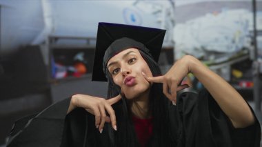 Mezuniyet üniformalı genç İspanyol kadın dışarıda bir şantiyede neşeli ifadeler sergiliyor, akademik başarıyı gerçek dünyadaki endüstriyel ortamlarla harmanlıyor..