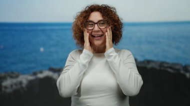 Kıvırcık esmer saçlı orta yaşlı kadın, arka planda engin mavi bir deniz ile deniz kenarında sürpriz ve neşe ifade ediyor..