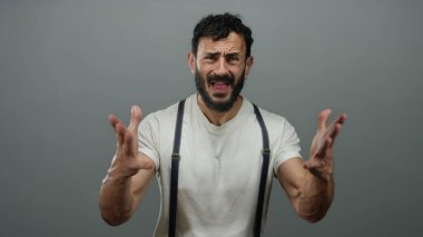 Sakallı İspanyol bir adam beyaz gömlekli, pantolon askısı takmış gri bir arka planda hayal kırıklığı yaşıyor..