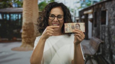 Gülümseyen kadın, Meksika pesolarını şehir caddesinde tutuyor, açık havada mutlu bir ifade ve neşeli tavırla para sergiliyor, orta yaşlı İspanyol kadını elinde parayla vurguluyor..