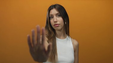 Beyaz kolsuz bluz giyen kendine güvenen İspanyol kadın stüdyoda turuncu duvarlı avuç içini ileri uzatıyor..