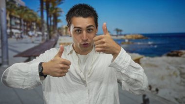 Beyaz gömlekli genç adam, arka planda palmiye ağaçları ve mavi okyanusla güneşli bir sahil gezintisine başparmak sallıyor, pozitifliğini ve dışarıda eğlenmeyi ifade ediyor..