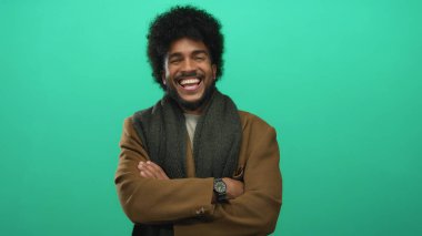 Yeşil bir arkaplanda kolları çaprazlanmış gülümseyen bir adam, kendine güveni ve sıcaklığı şık kahverengi bir ceket ve siyah bir atkıyla yayılıyor..