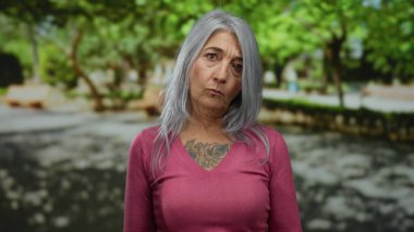 Gri saçlı, pembe kazaklı yaşlı bir kadın güneşli bir parkta dikiliyor. Yeşillik ve banklarla çevrili doğal ifadesini sergiliyor..