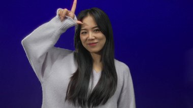 Genç Çinli kadın, kaybedenleri işaret eder. İzole edilmiş mavi arka planda. Sıradan bir süveterle gülümsüyor..