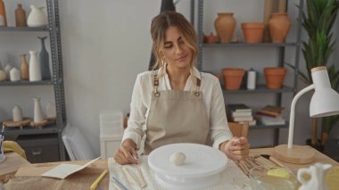 Sarışın genç bir kadın, zanaatkar stüdyosunda kapşonlu önlük giyerek çömlek tekerinde oturur. Sabır üzerine kafa yorar..