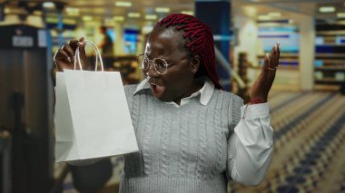Gözlüklü ve süveterli bir kadın alışveriş torbası tutuyor. Arka planda bulanıklık var. Modern bir mağaza ya da alışveriş merkezi ortamı öneriyor..