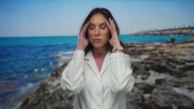 Deniz kenarında gözleri kapalı, elleri şakakların üzerinde beyaz bir ceket giymiş bir kadın okyanus kıyısındaki parlak mavi gökyüzünün altında baş ağrısını yakalıyor..