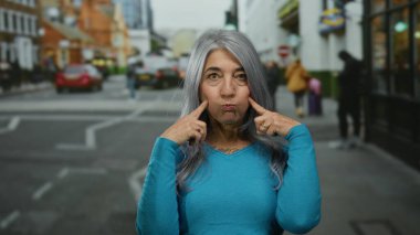 Yoğun bir şehir sokağında, arka planda arabaları ve yayaları olan sokak hayatını gözler önüne seren, neşeli bir ifade sergileyen gri saçlı son sınıf kadını..