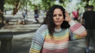 Renkli kazaklı kıvırcık saçlı kadın, güneşli parkta ağaçların bulanık arka planıyla ve yürüyen insanlarla ilgili jest yapıyor..
